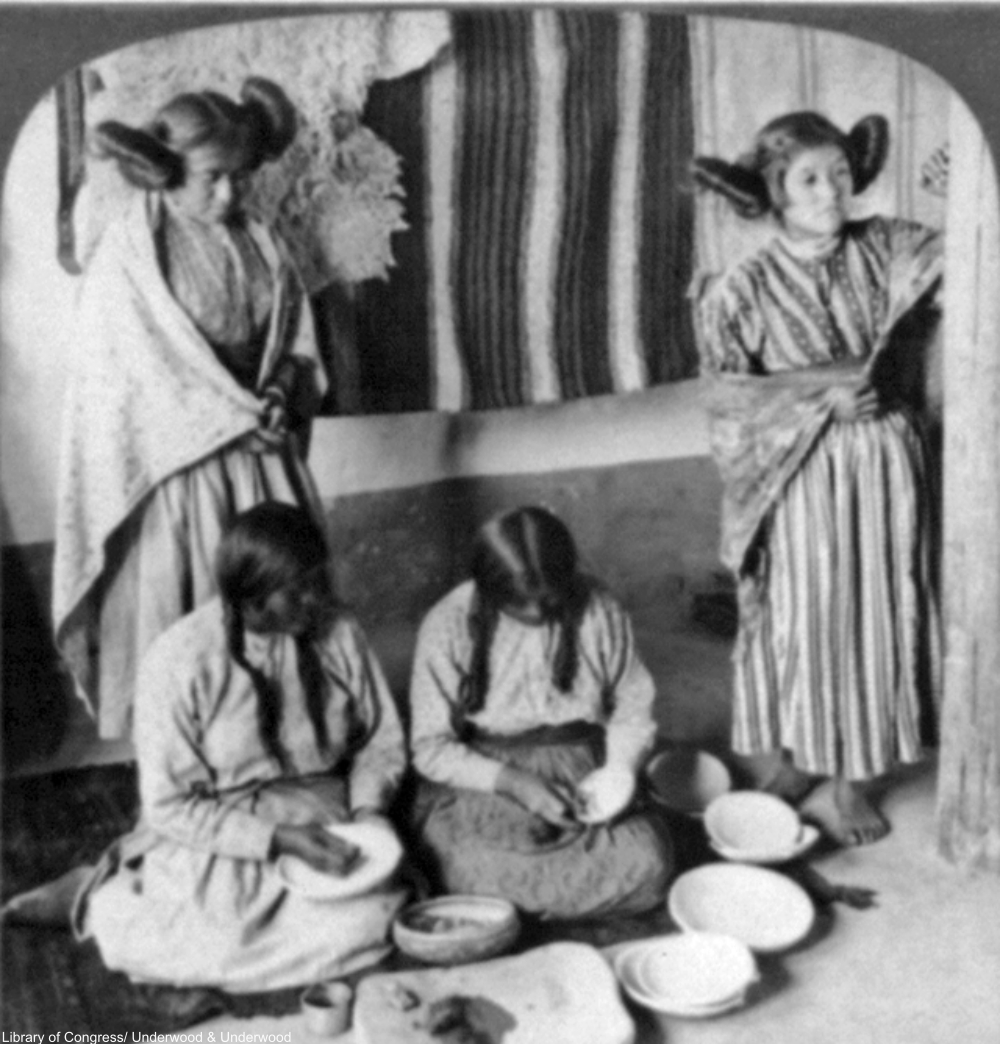 Four Hopi women, two are decorating pottery and two, with squash-blossom hairstyles, are standing in the background, circa 1903